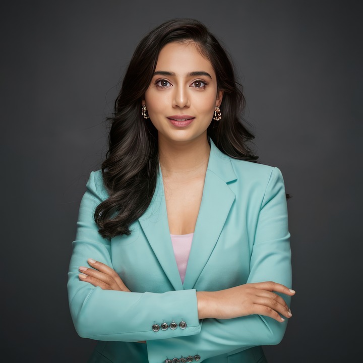A professional Indian woman with long, dark hair, wearing a mint green blazer over a pink top, smiles with arms crossed against a grey background.